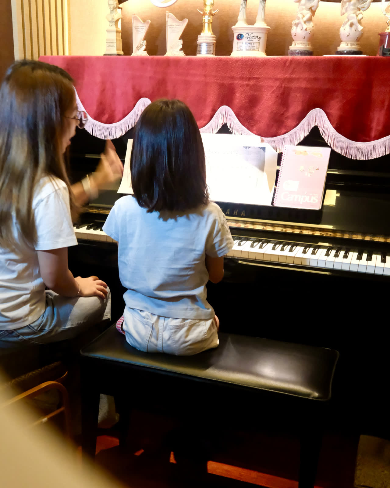 A lesson in progress: a teacher and a young student side by side at the upright piano, seen from behind, an open score on the desk.