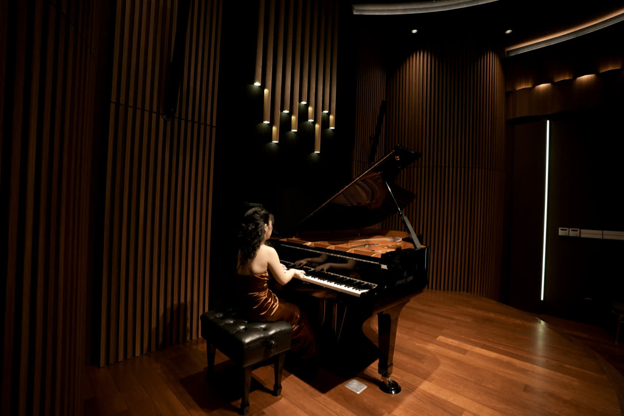 The studio: a grand piano with its lid open on a timber floor, a pianist at the keyboard, the walls lined with vertical acoustic slats.