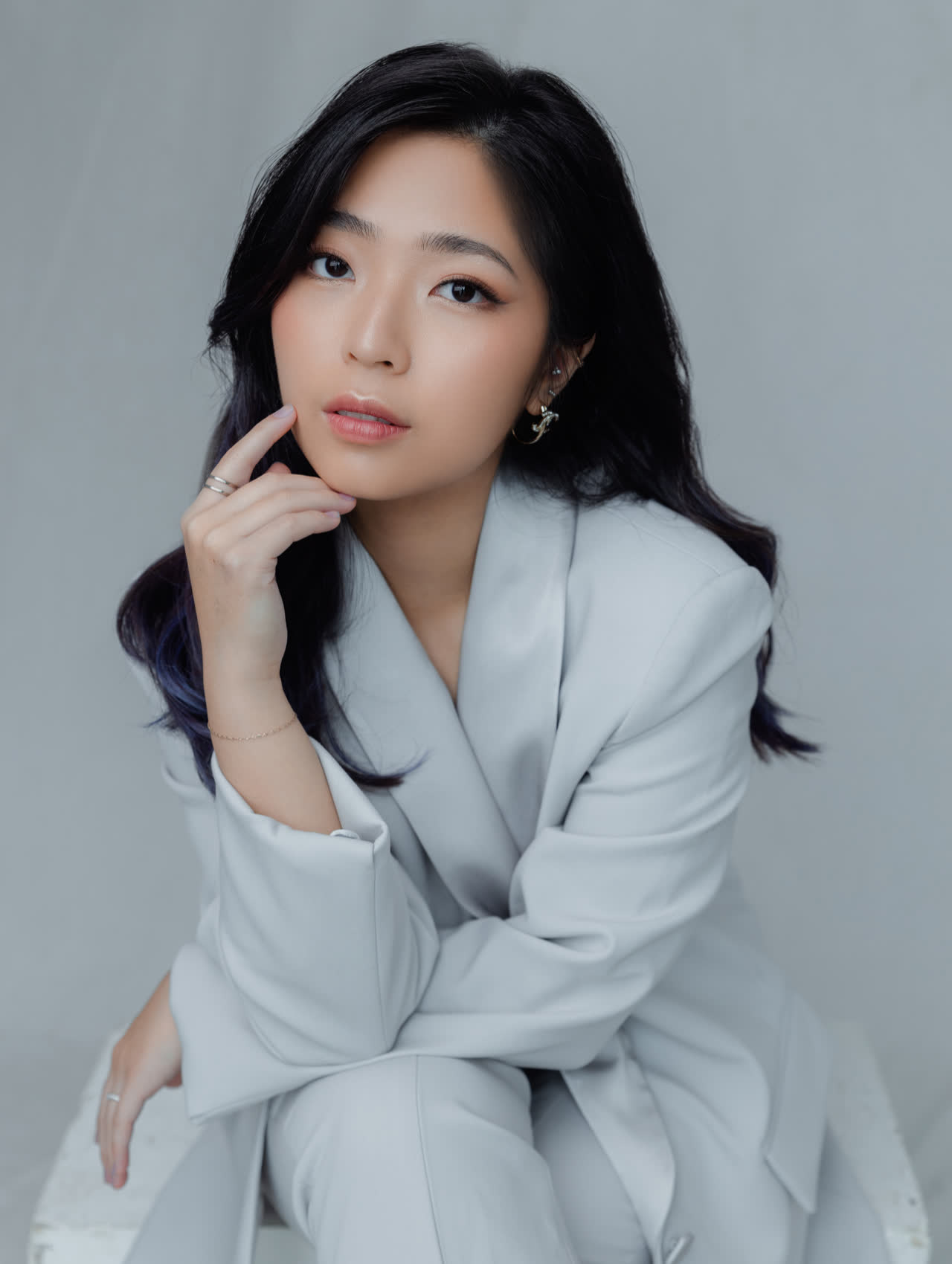 Sally Yapto, seated, in a pale grey suit against a soft grey studio backdrop.