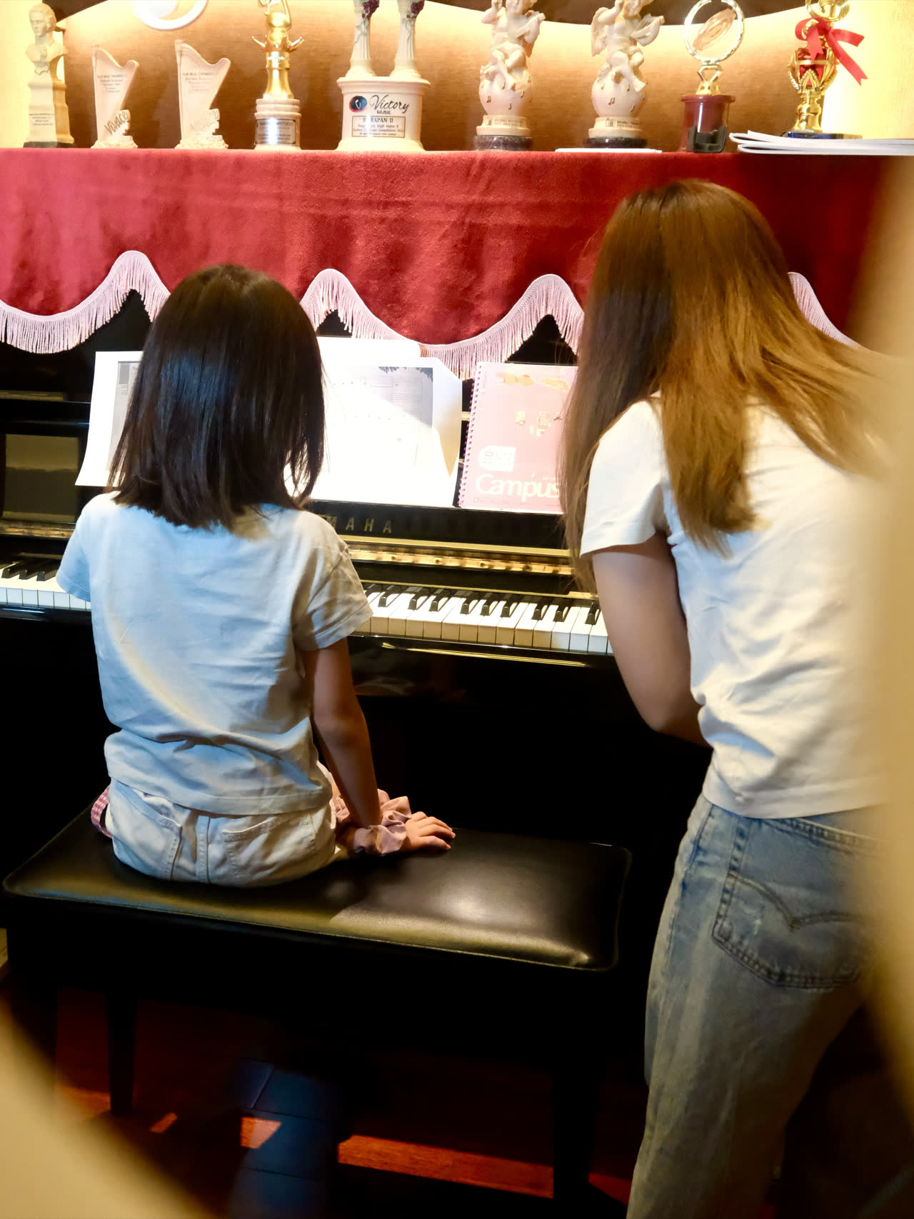A lesson in progress: a young student at the upright piano with an open score, the teacher standing beside her, watching her hands.