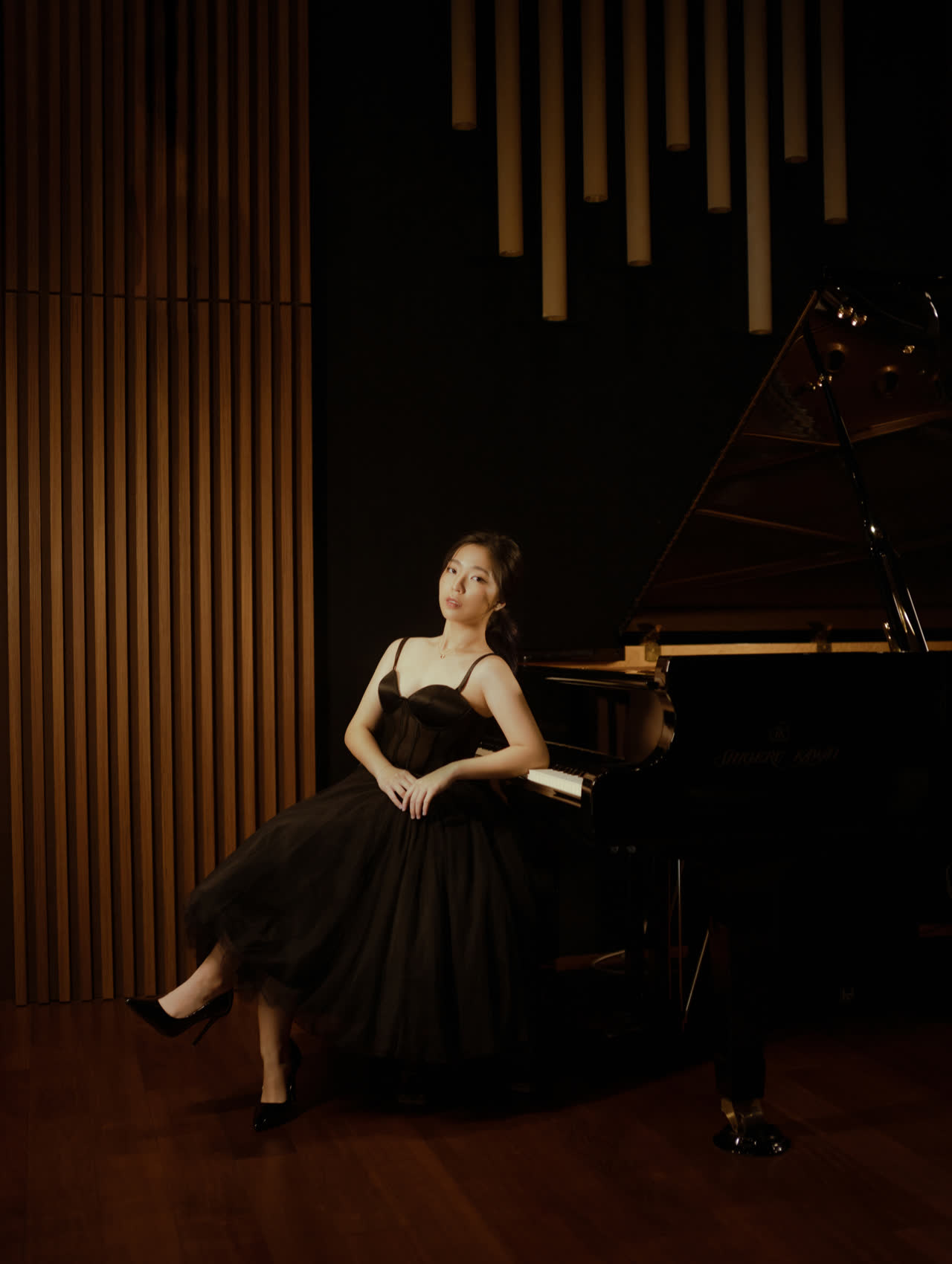 Sally Yapto seated at the grand piano in the studio, in a full black gown, the timber slat wall behind her.