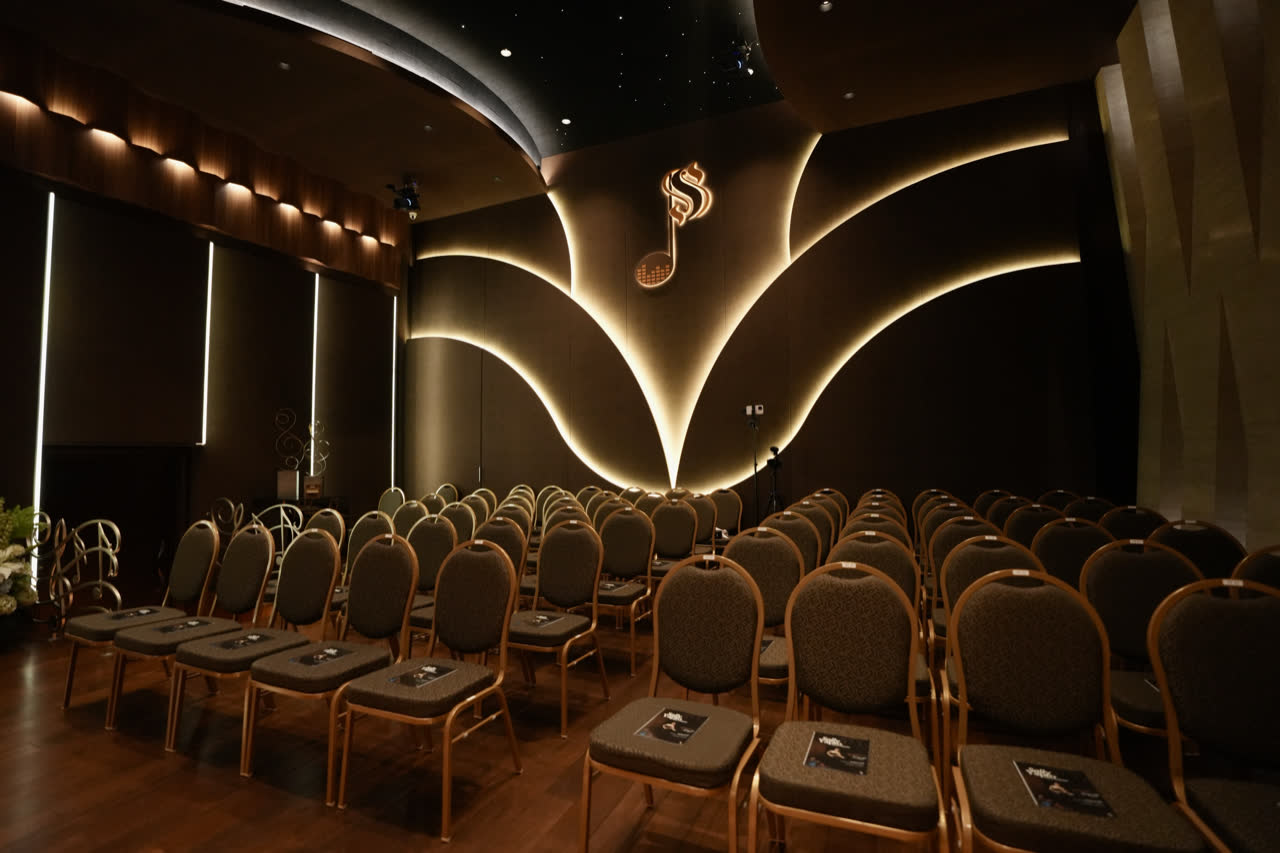 Kursi aula dilihat dari samping, menghadap logo not seperdelapan Symphony yang disinari dari belakang, dengan penyangga mikrofon dan kamera tetap terpasang.