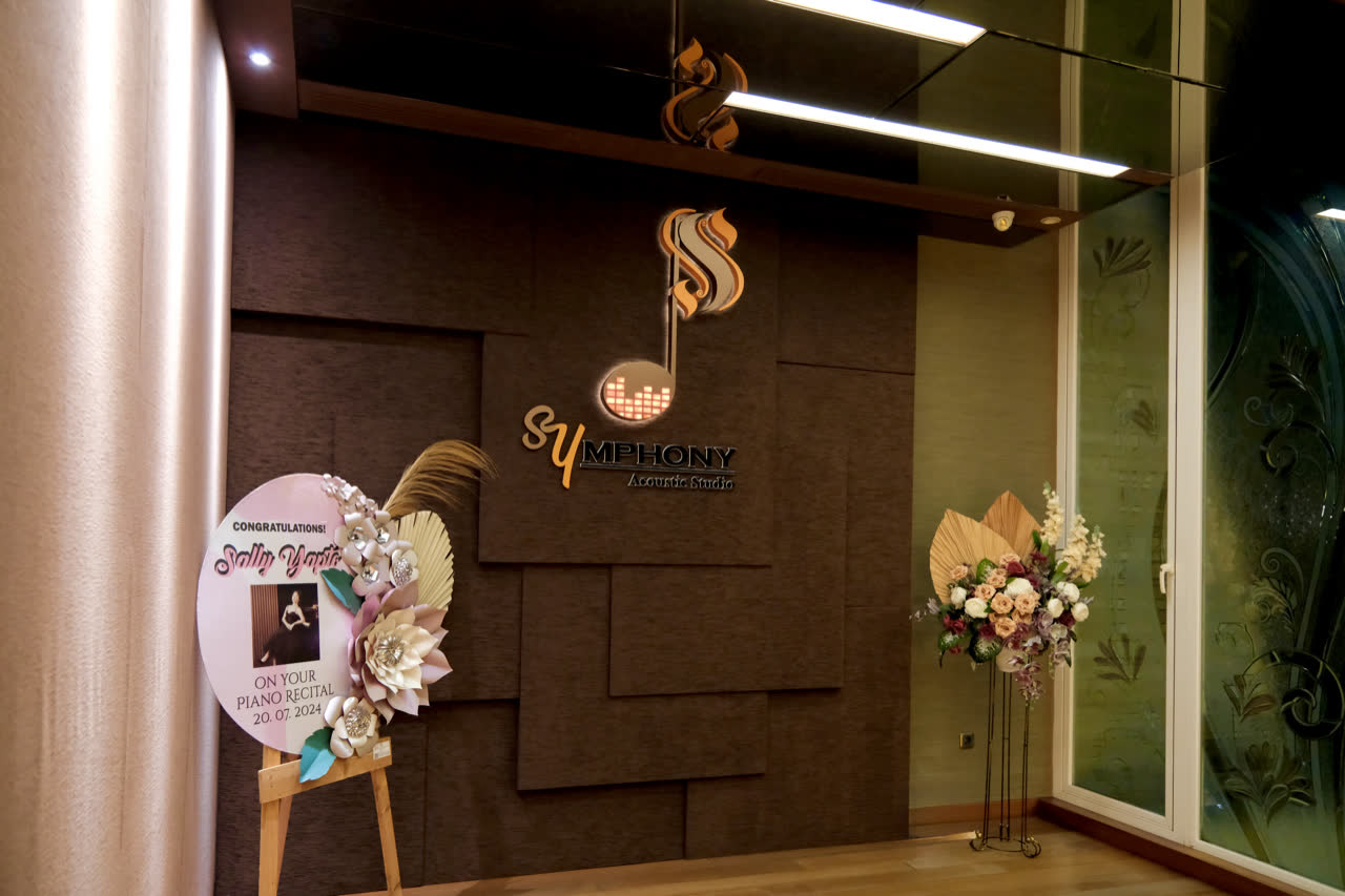 Pintu masuk studio: logo Symphony Acoustic Studio yang menyala di dinding berpanel di samping pintu kaca.