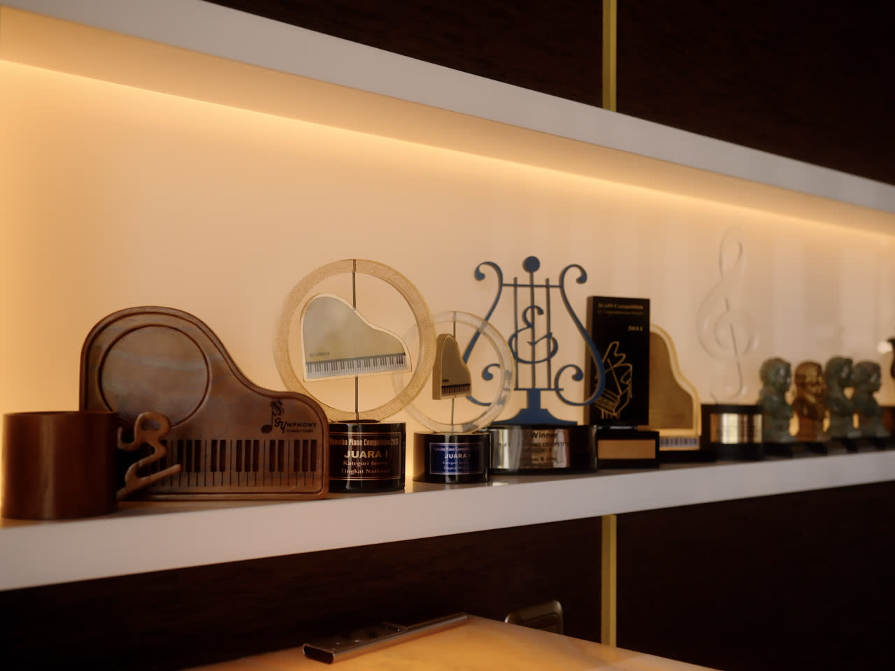 Rak berlampu di lounge berisi piala kompetisi piano dan patung-patung kecil bertema musik.